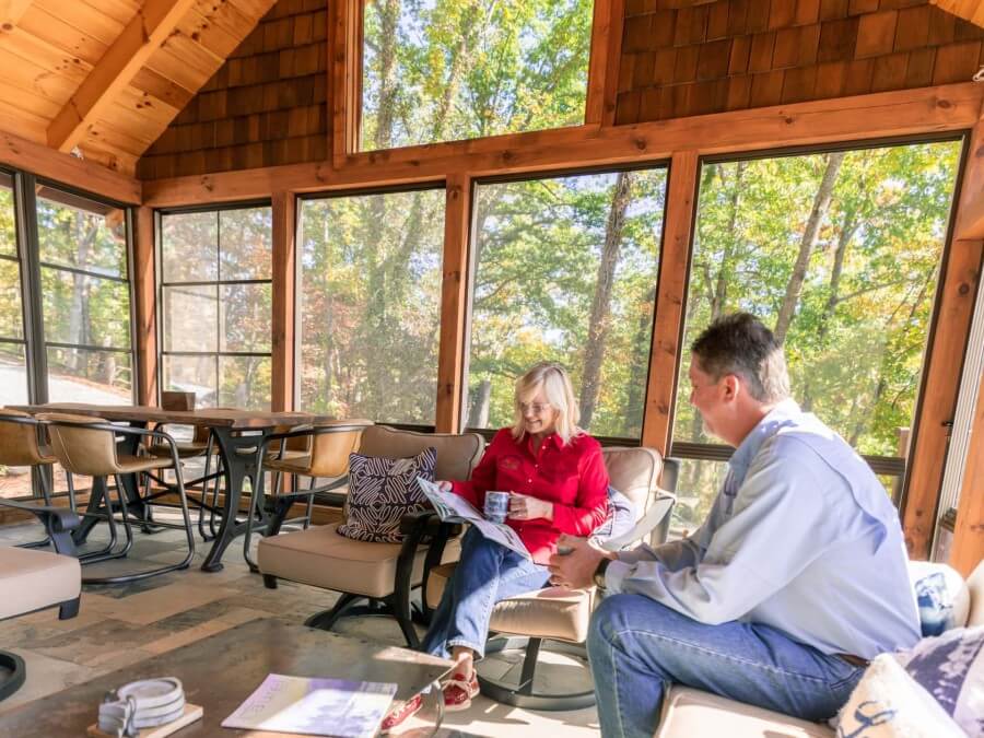 Husband and wife enjoy their coffee and a read in a wooden three season room, filled with windows and surrounded by trees.