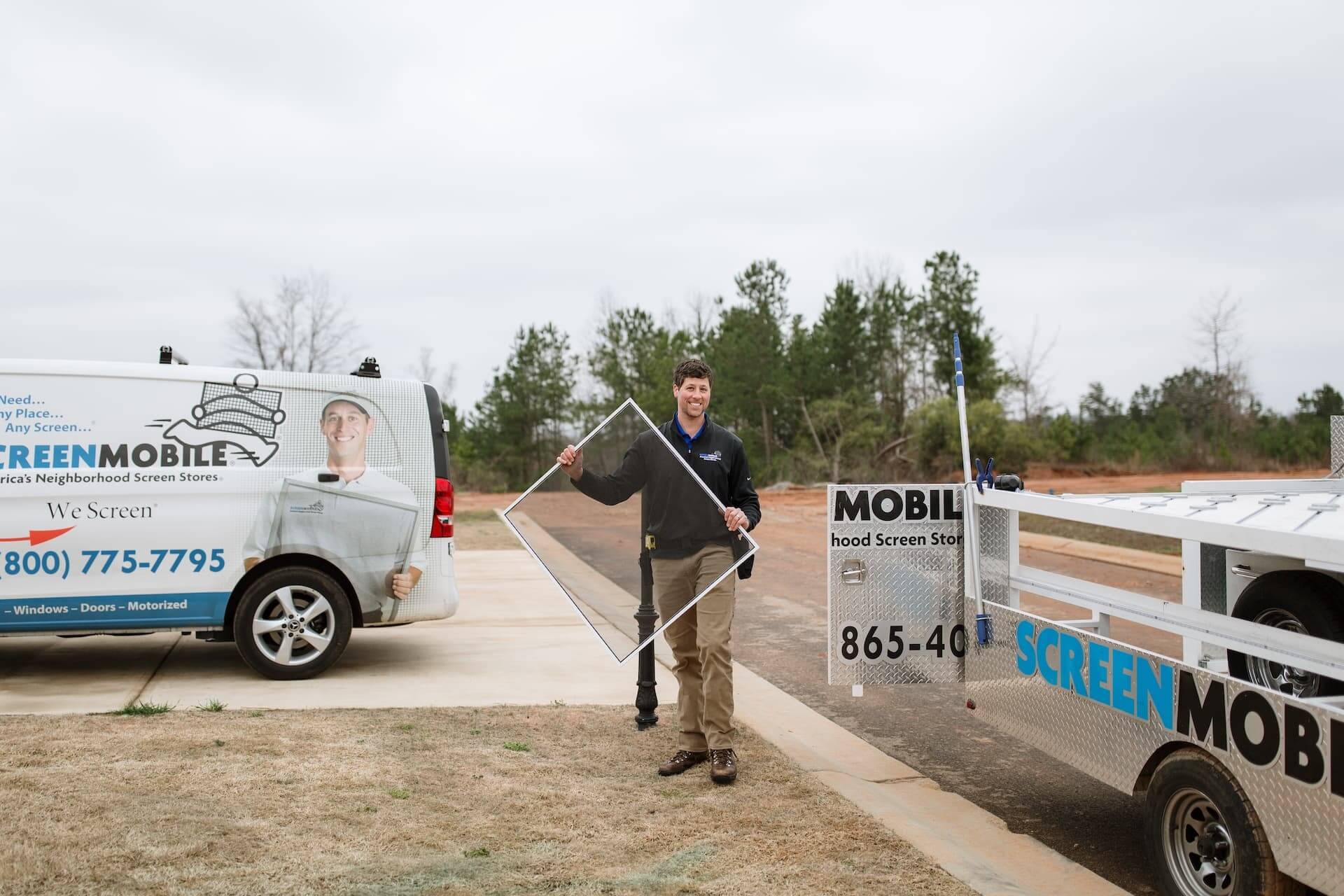 A Screenmobile pro carries a screen to a client's home. A Screenmobile van and trailer are parked at the home.