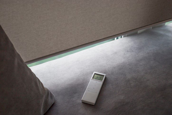 A remote control for motorized window shades is lying on a carpeted floor, indicating a modern home automation setup.