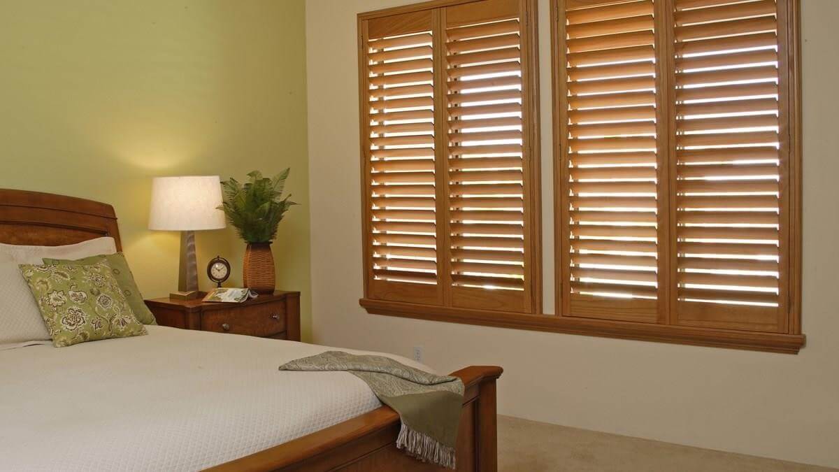 Norman Window Fashions - wood finished shutters in a bedroom with a white bed